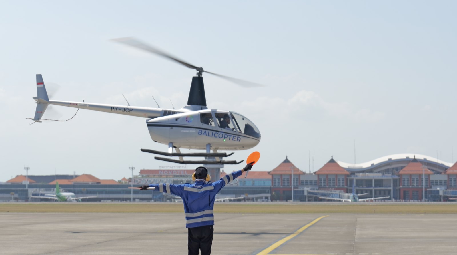 A Balicopter helicopter hovering at Ngurah Rai International Airport, guided in by a marshaller